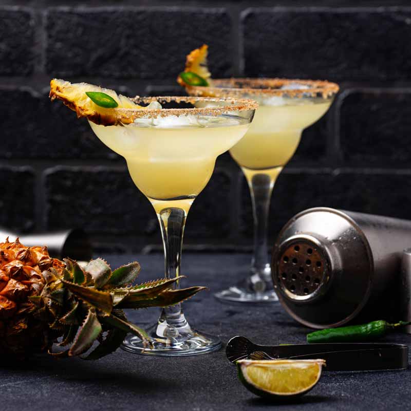 Two cocktails in margarita glasses with garnishes.