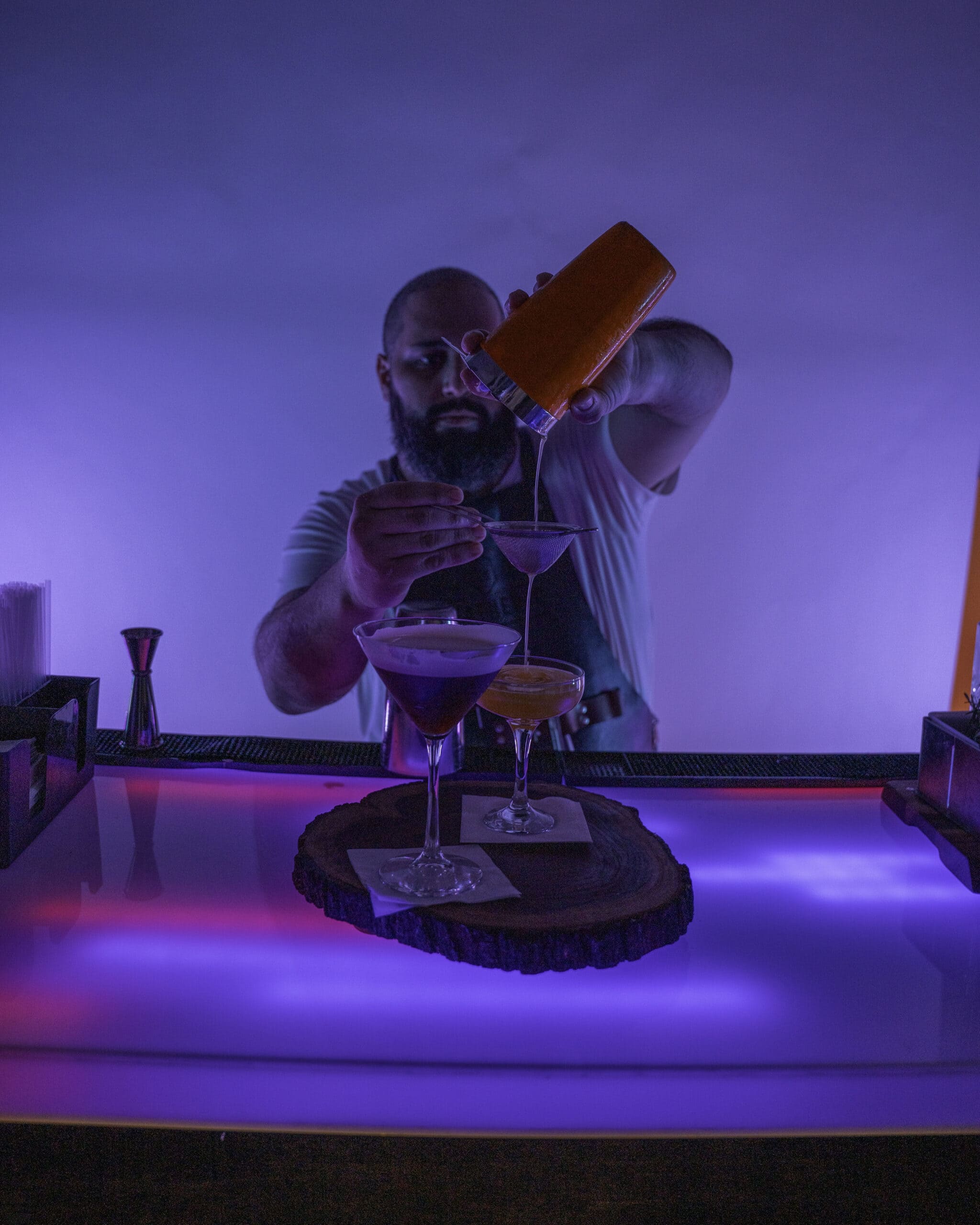 Bartender pouring a cocktail into a glass behind a bar.
