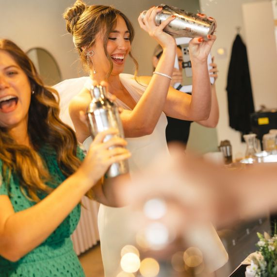 Two women smiling and shaking cocktail shakers in a bright indoor setting, enjoying a lively drink-making moment.