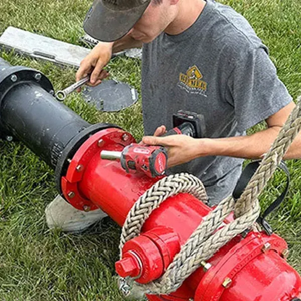 A plumber from Beemer Plumbing is installing a new tankless hot water heater in a laundry room in Spring Hill, KS. The image shows the technician carefully mounting the compact gas-fired tankless unit on the wall and connecting the copper pipes, PEX lines, and venting system. The surrounding area features laundry appliances and professional tools, capturing the professional installation process in a local residential basement.