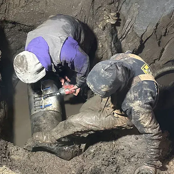 A Beemer Plumbing team member installs a new underground water line at a residential property in Spring Hill, KS. A technician in a branded blue shirt carefully connects the grey polyethylene pipe and fittings, while a colleague manages the pipe coil next to a trench. A Beemer Plumbing service truck is parked in the driveway of the high-end home in the background, showcasing the company's professional water line repair and replacement services.
