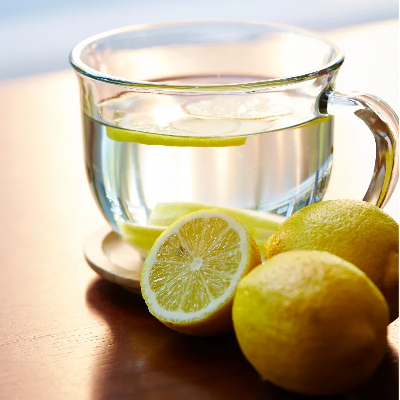 Glass cup of water with fresh lemon slices beside lemons, evoking refreshment, energy, and soothing nourishment for busy moms