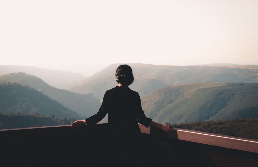 A woman standing peacefully on a balcony overlooking a vast mountain range, representing the clarity and grounded strength found through intuition during a challenging cancer season.