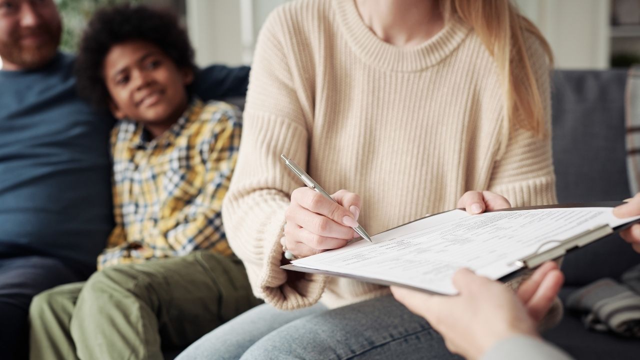 A close-up of a mother’s hand holding a pen over a registration clipboard, with her family in the background, representing the decision-making process for kids' sports and extracurricular activities.