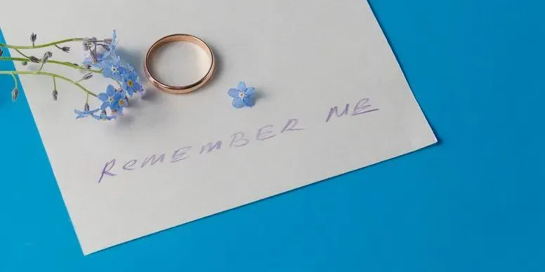 A pair of wedding rings placed on a white envelope with a flower, representing obituary writing assistance.