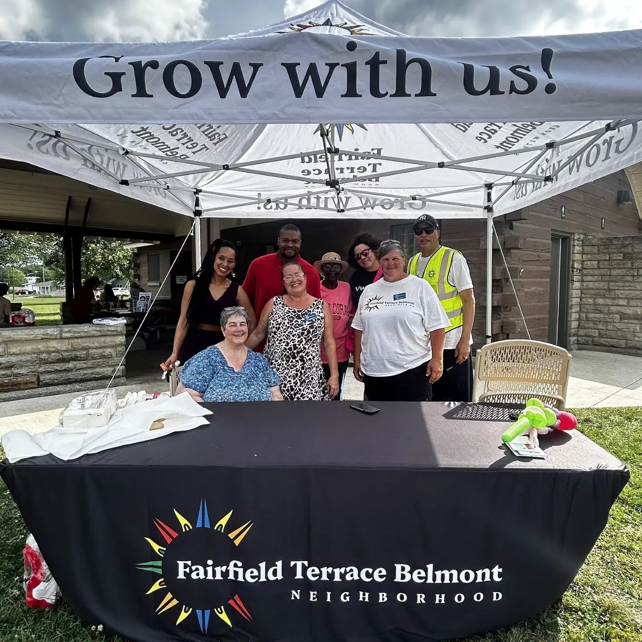 A varied group of neighbors sit and stand smiling behind a table with a black stretch tablecloth that says "Fairfield Terrace Belmont Neighborhood" and has a multicoloured sun around the word Fairfield. They stand under a white pop-up tent that says "Grow with us!" and behind them is a wood and stone building and a covered area with seating.