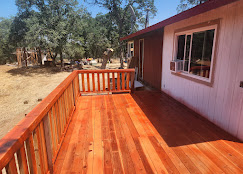 A newly stained redwood deck with a matching railing attached to a white and brown house under a clear blue sky.