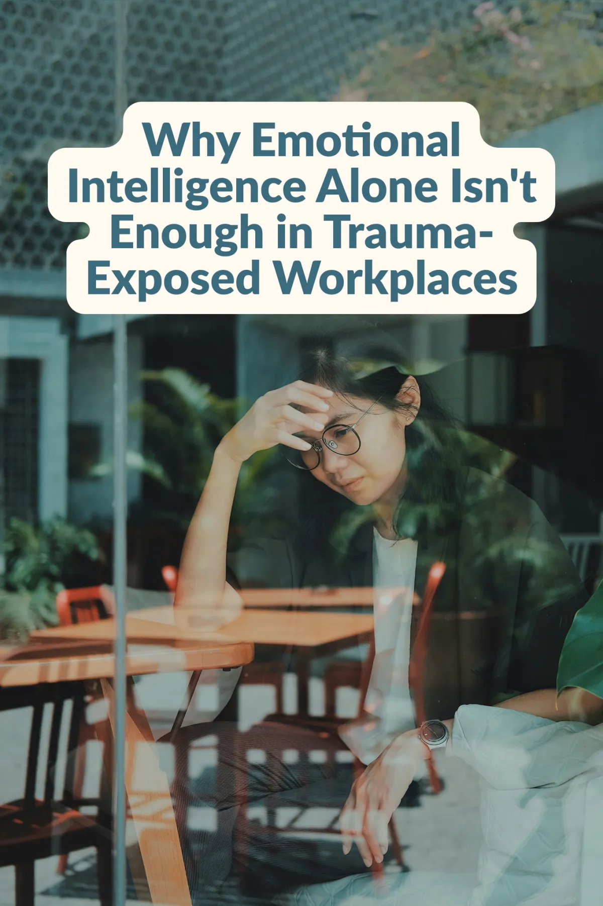 A woman wearing glasses rests her hand on her forehead looking stressed or fatigued, photographed through a window with greenery and café furniture visible in the background. Overlaid text reads: 'Why Emotional Intelligence Alone Isn't Enough in Trauma-Exposed Workplaces.'