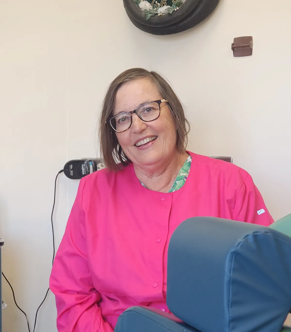 Dr. Anne McDonald, wearing a pink coat, sits in her dental office chair, smiling warmly at the camera.