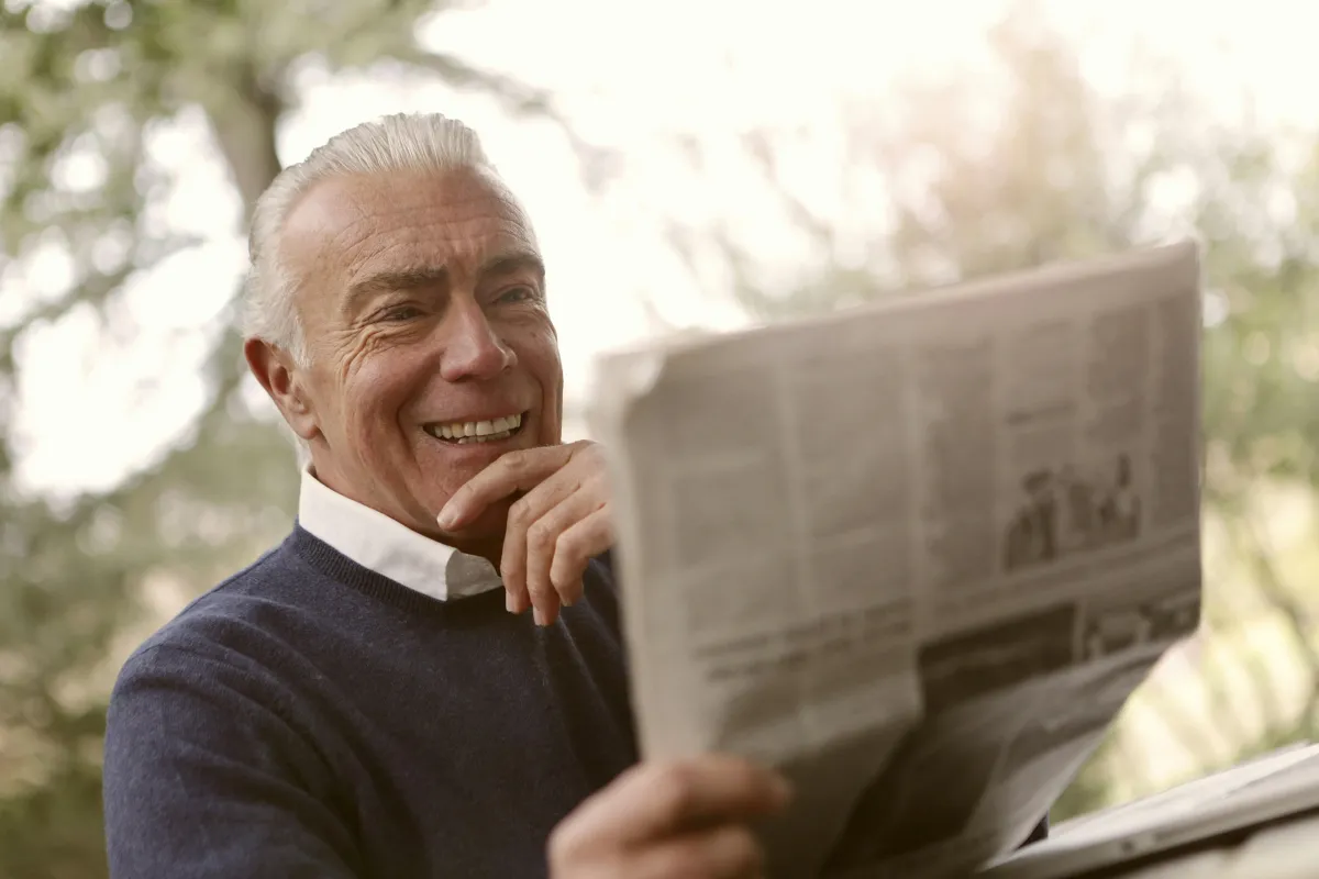 Man smiling while reading the newspaper outdoors.