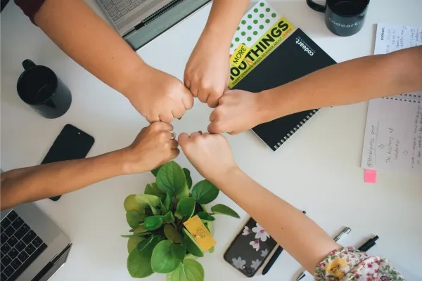 An overhead view of five people fist-bumping in a circle above a white desk, which is scattered with laptops, smartphones, black mugs, notebooks with "codeart" branding, a book titled "MORE 100 THINGS", and a potted green plant.