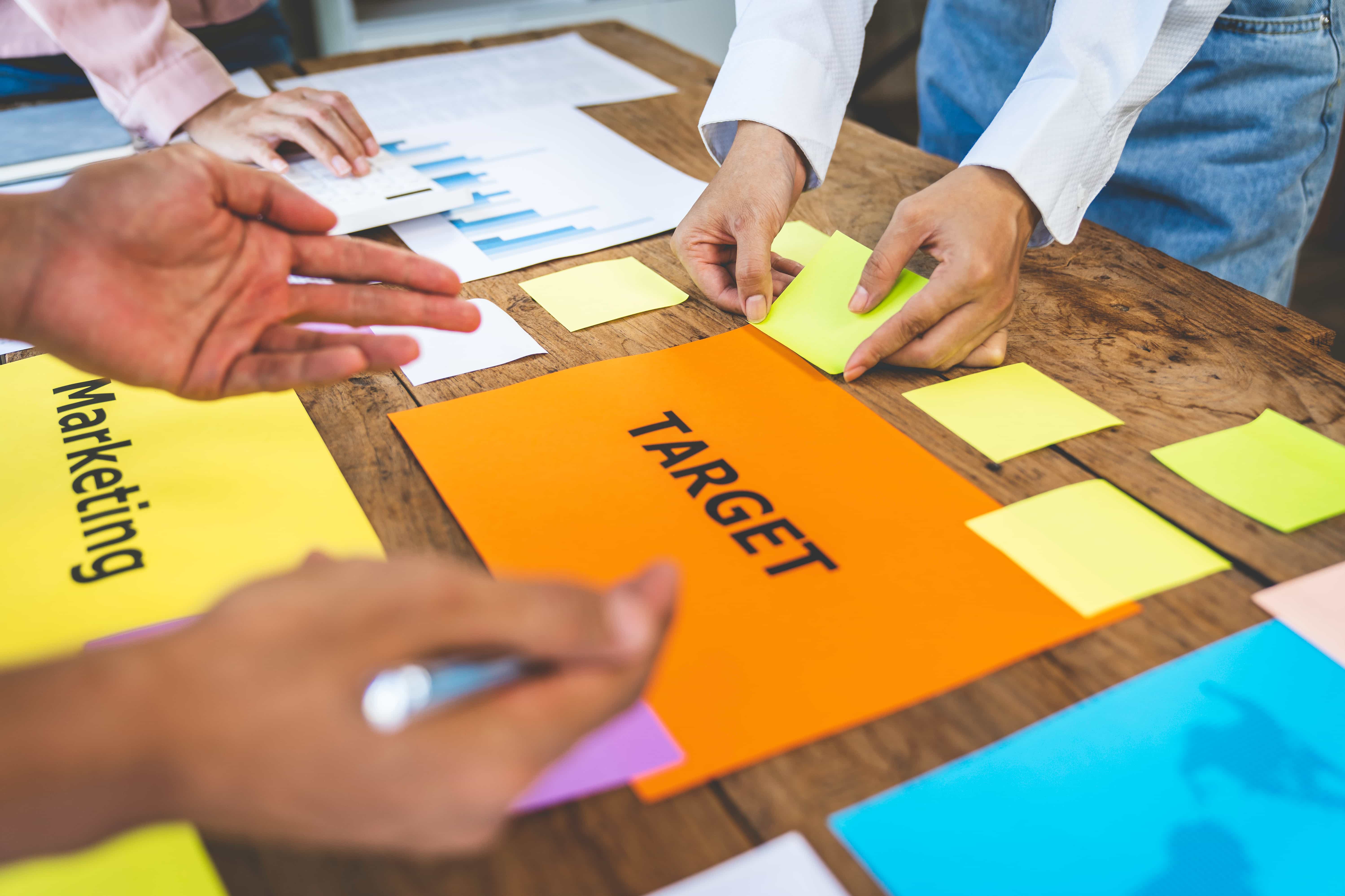 A collaborative team in Austin, TX, working around a wooden table with colorful sticky notes and a large orange sign labeled "TARGET" to define marketing goals.