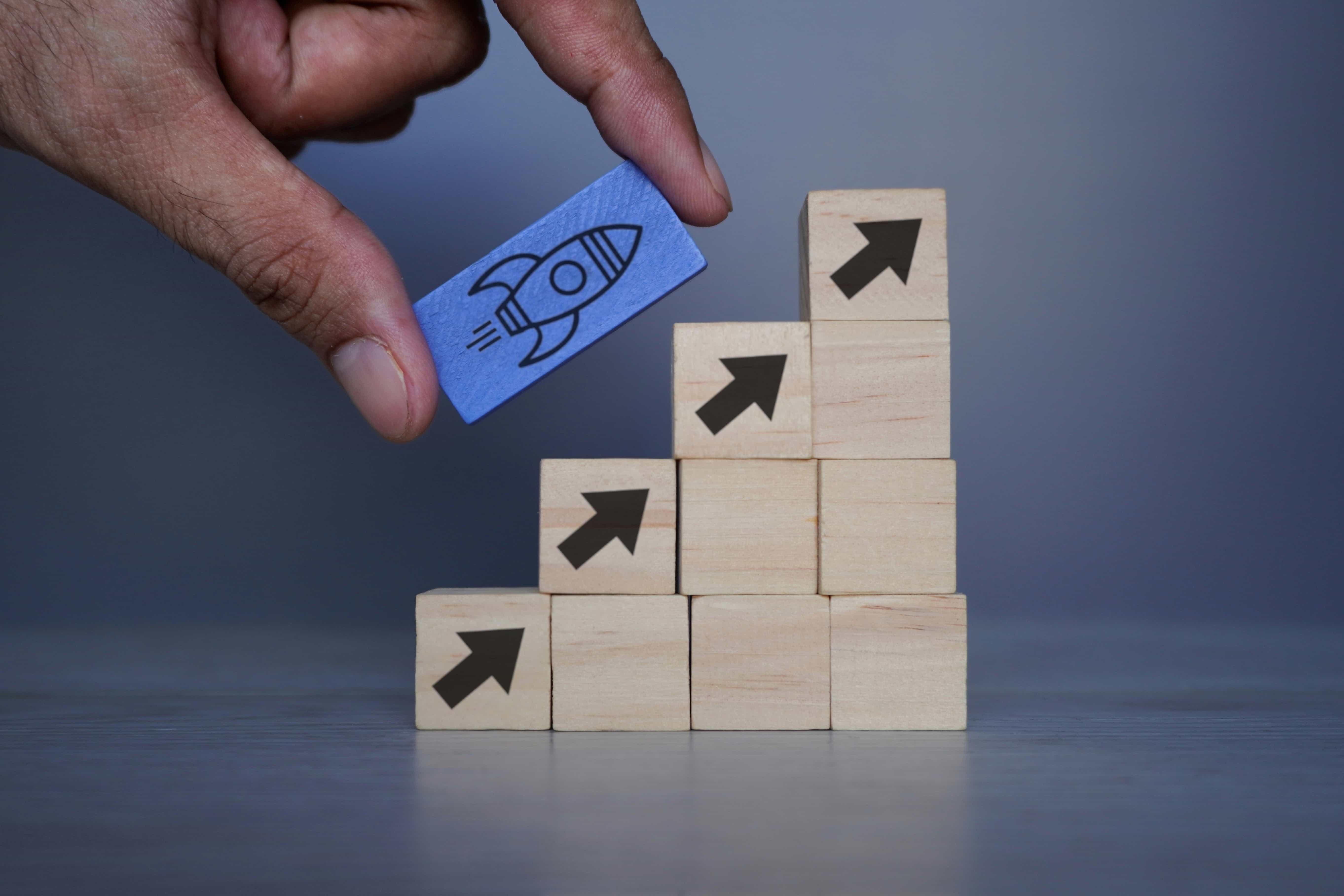 A hand placing a blue block with a rocket icon on top of a rising wooden block staircase with upward arrows, representing scaling conversions through a fast response speed-to-lead framework.