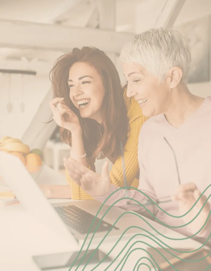 Two smiling women, one younger and one older, laughing together while looking at a laptop during career coaching session