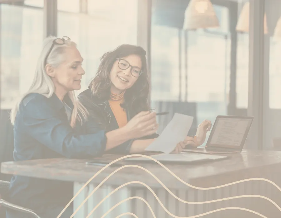 Two professional women collaborating at a table with a laptop and documents, representing career coaching, mentorship, and goal-setting.