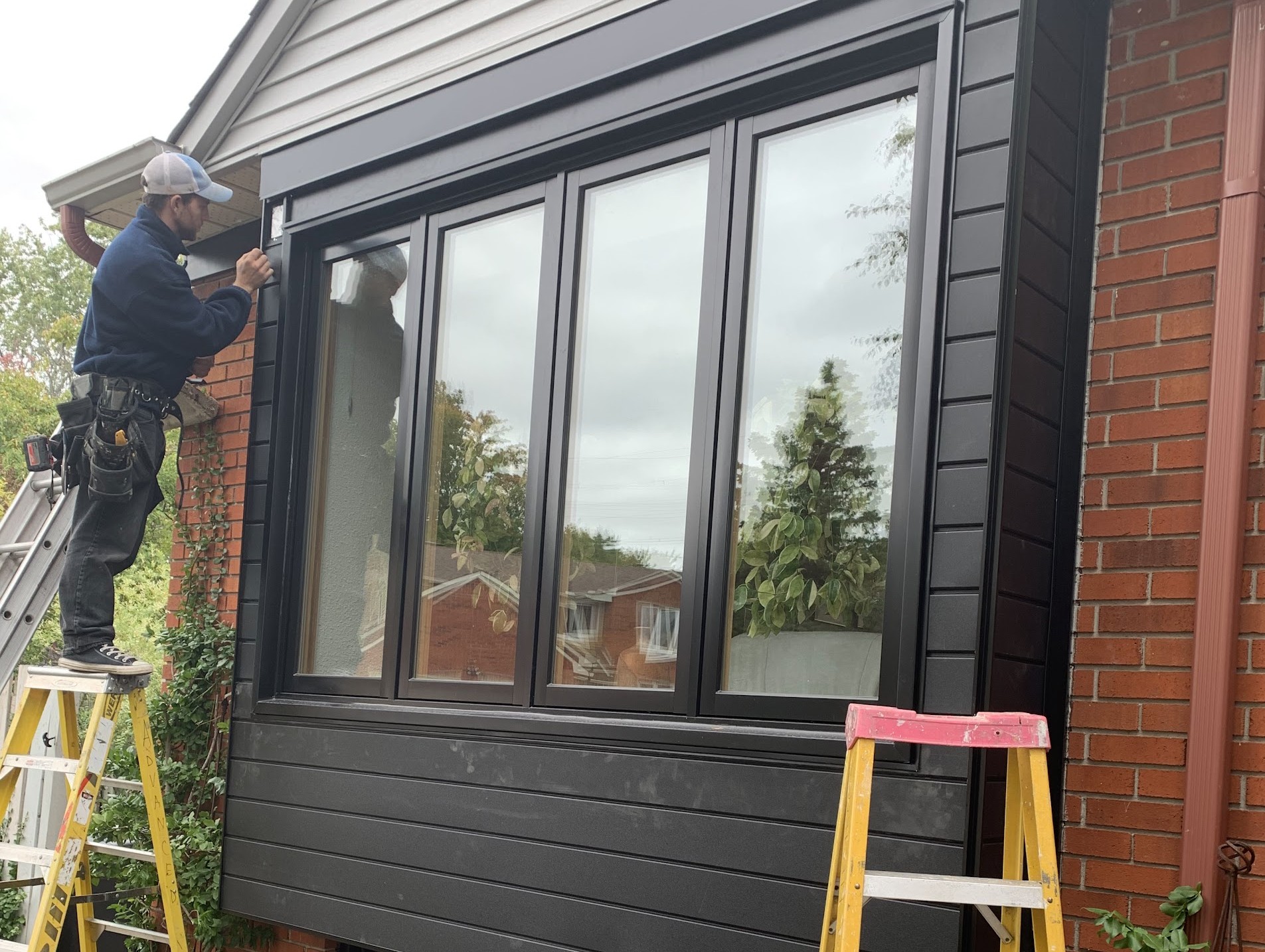 A professional installer finishing the exterior trim of a large, modern four-pane black window unit with matte black siding on a red brick house.