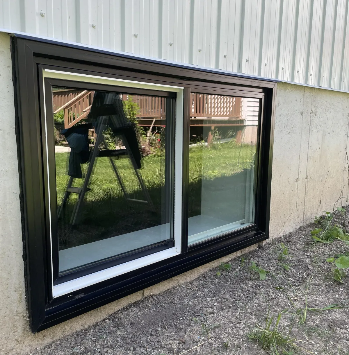 A freshly installed, modern egress window with a sleek black frame is set into a concrete foundation wall beneath white metal siding. Bright green grass and a wooden deck are reflected in the clear glass, with dirt and gravel ground in the foreground.
