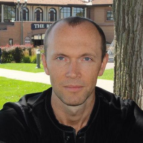 professional man sitting outdoors near a tree with a building and green lawn in the background