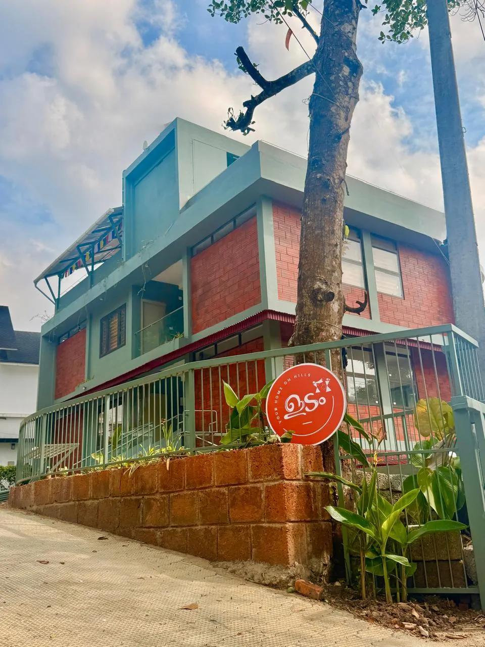 A candid photo of a smiling local family standing in front of a traditional homestay house, surrounded by flowering plants and greenery. The family wears casual attire, radiating warmth and hospitality in a welcoming outdoor setting.