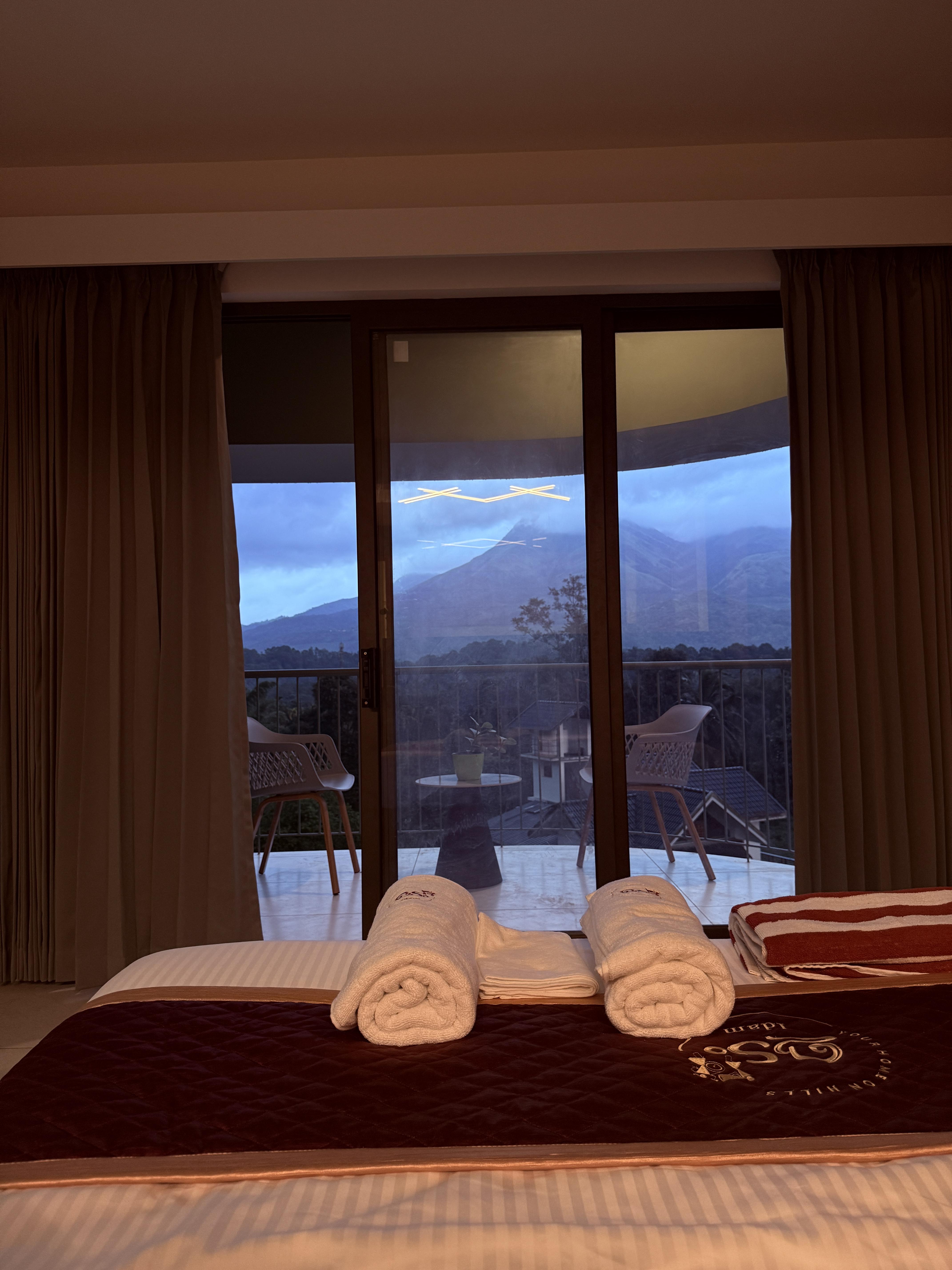 A misty morning view from the homestay balcony, overlooking rolling green hills and a winding river. The landscape is bathed in soft golden light, with wildflowers blooming along the wooden railing.