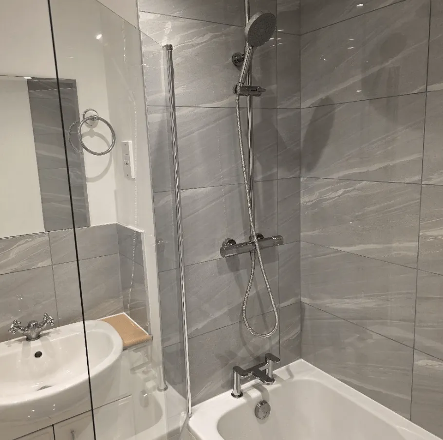 Grey tiled bathroom with shower over bath and basin by Willis Building Services London