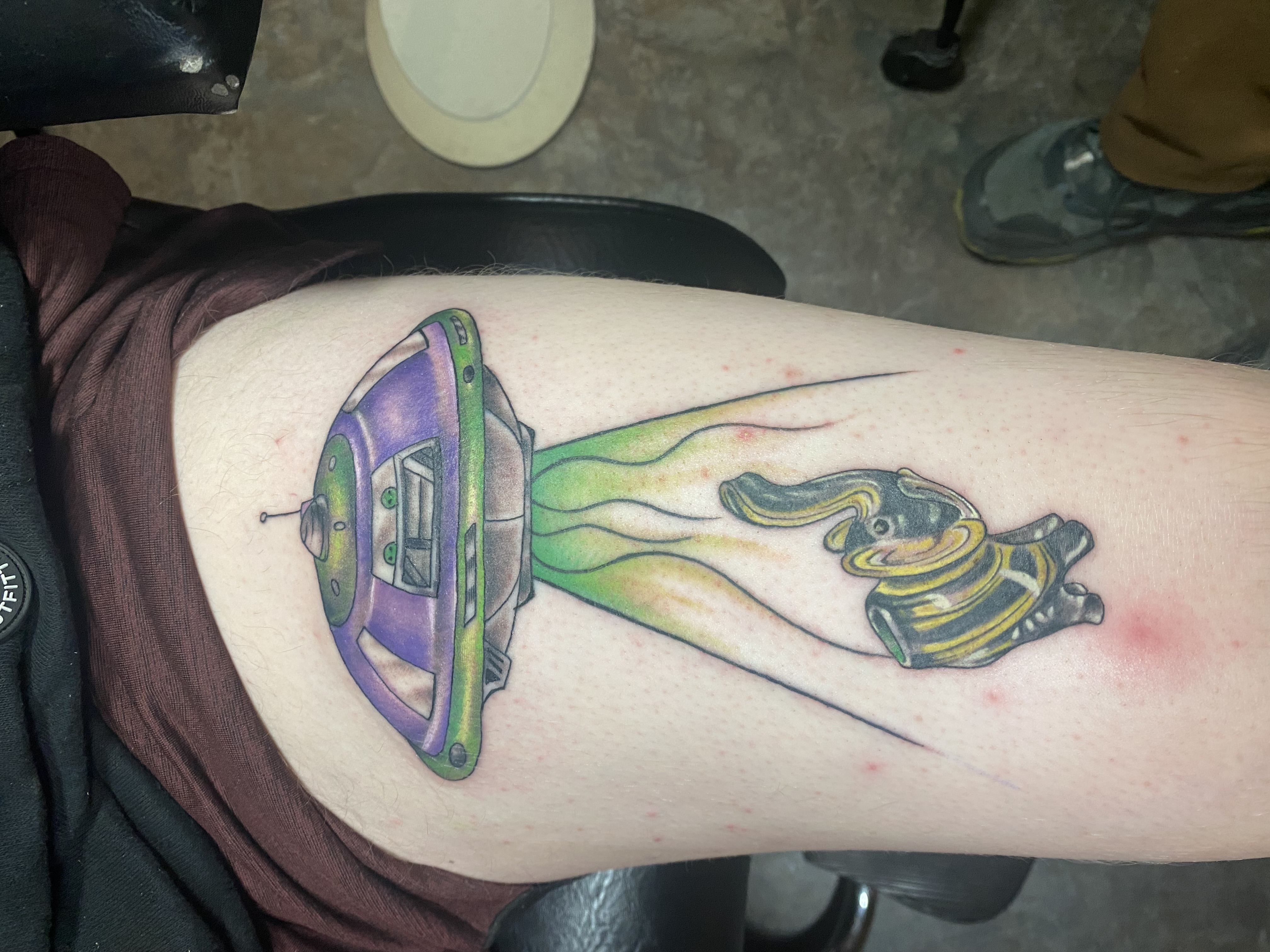 Minimalist linework tattoo of a botanical motif on a man's ankle, captured in a bright, airy space with a neutral backdrop. The style is clean and contemporary. 1:1 aspect ratio.