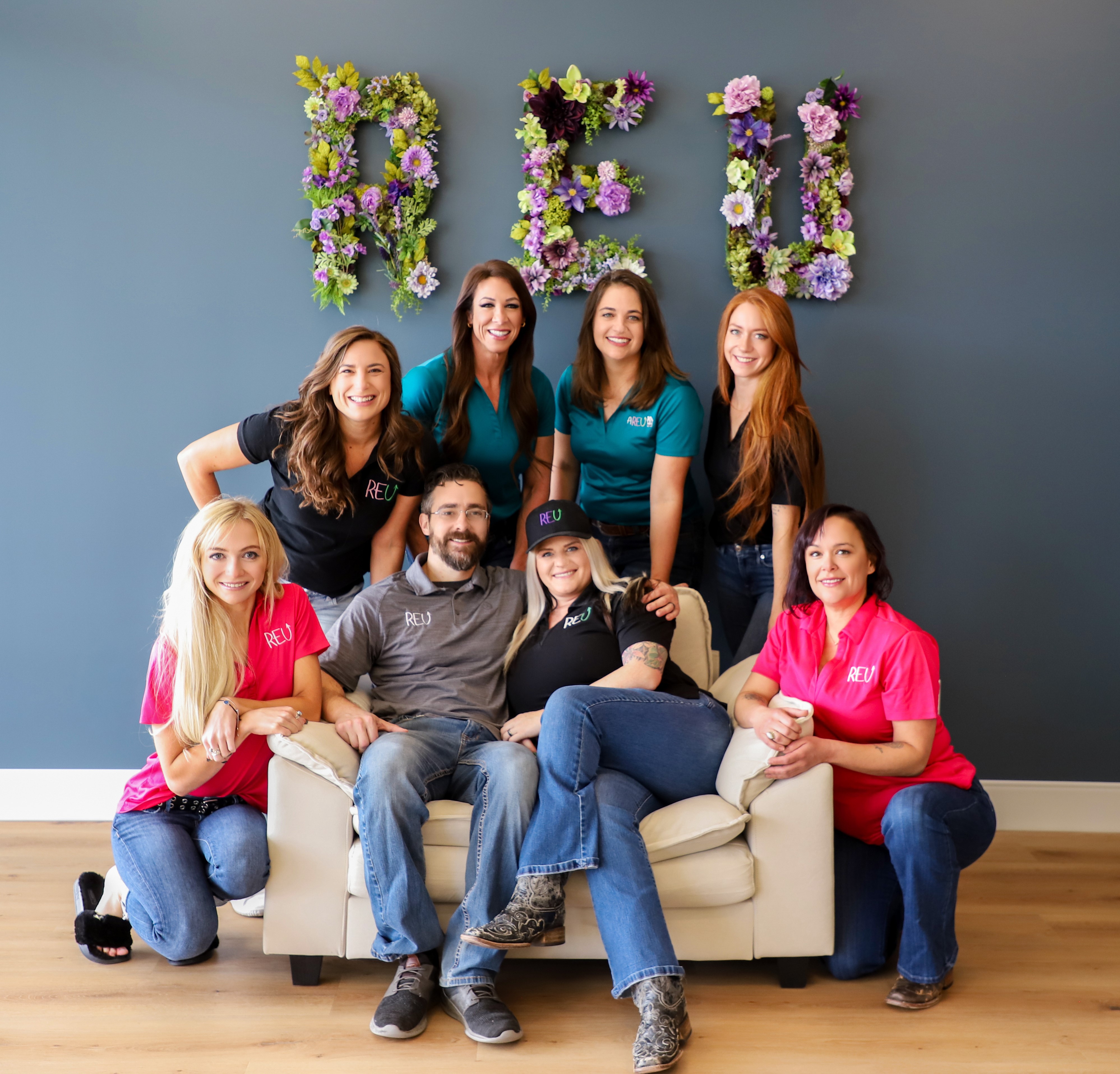 A diverse team of REU professionals standing in front of a local home, smiling, wearing branded shirts, with tools and design plans in hand, suburban neighborhood in background, bright daylight.