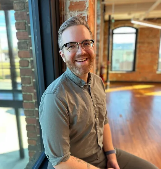 A portrait of a friendly, bearded man in his 40s, wearing a casual shirt, standing in front of a bookshelf filled with creative books and art objects.