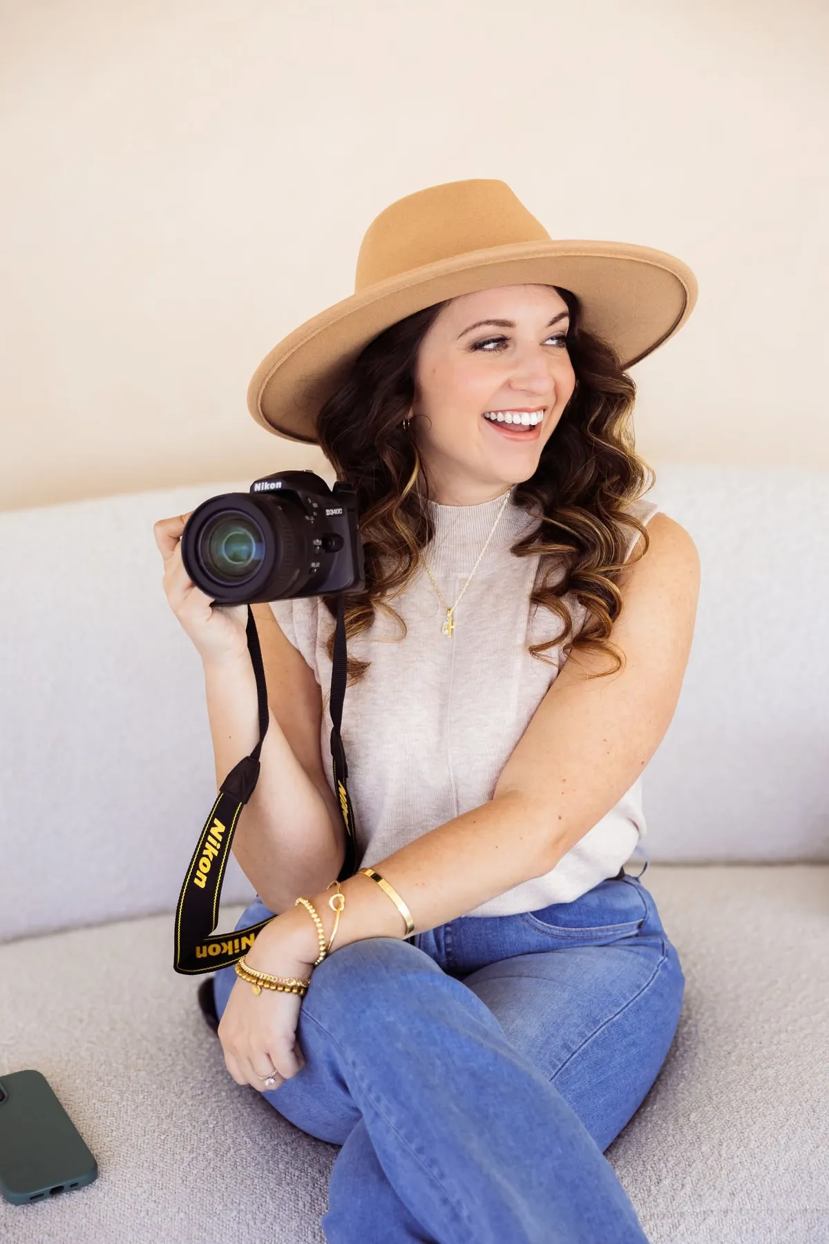 Woman in pink blazer sitting in a chair, holding phone out for a selfie in a minimal white room with a small plant.