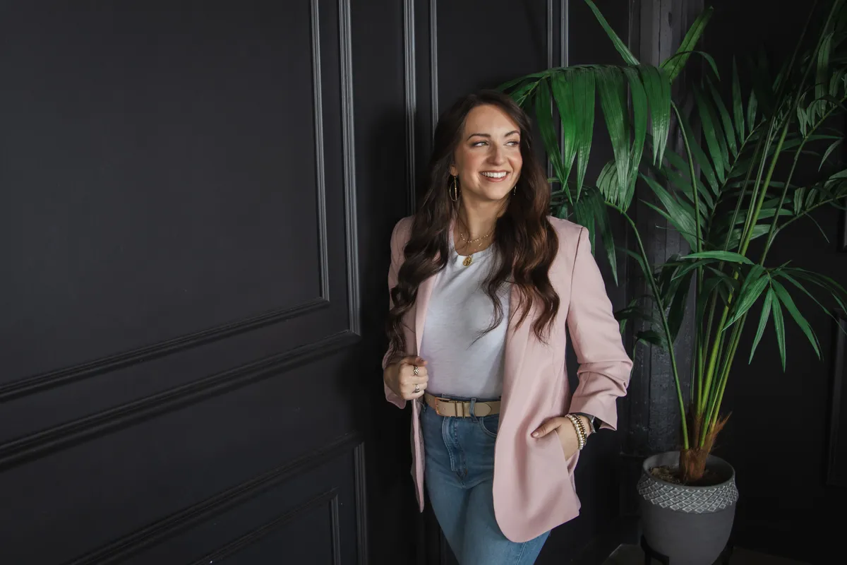 Portrait of a smiling woman wearing a pink blazer, white shirt, and blue jeans, standing indoors next to a dark gray paneled wall and a potted green plant