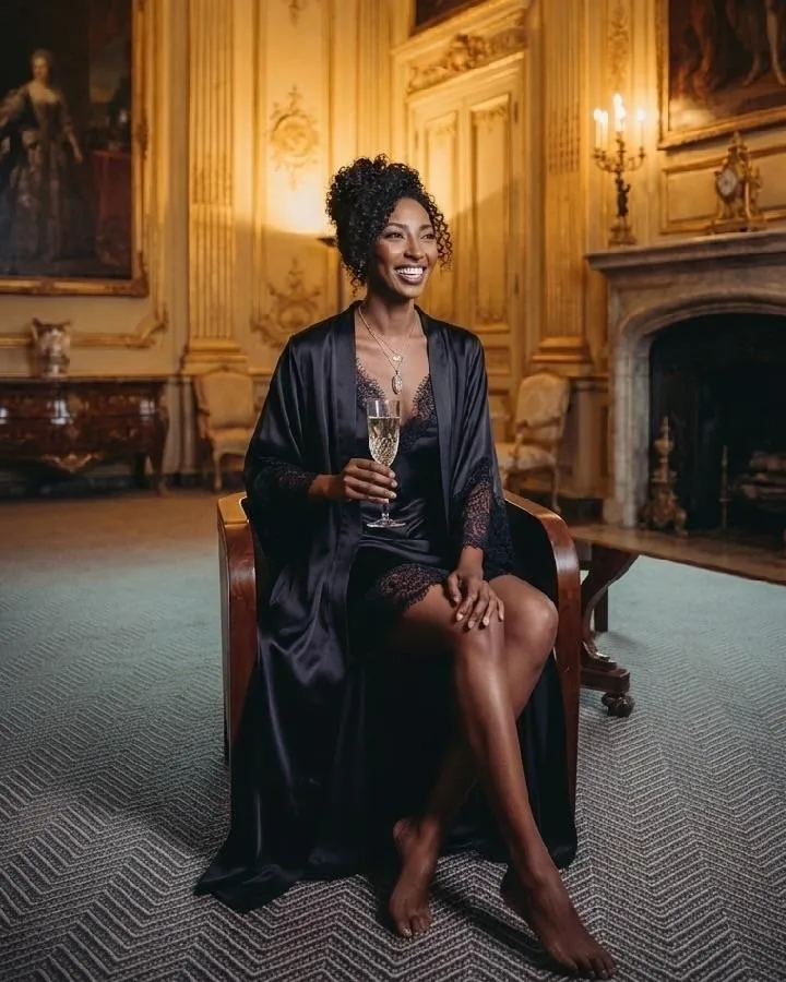 Woman wearing a dark robe and lace outfit sitting in a chair, holding a glass, inside a classic interior with a fireplace, artwork, and decorative wall details.
