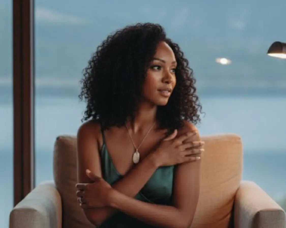 Woman with curly hair sitting in a chair near a window with water in the background.