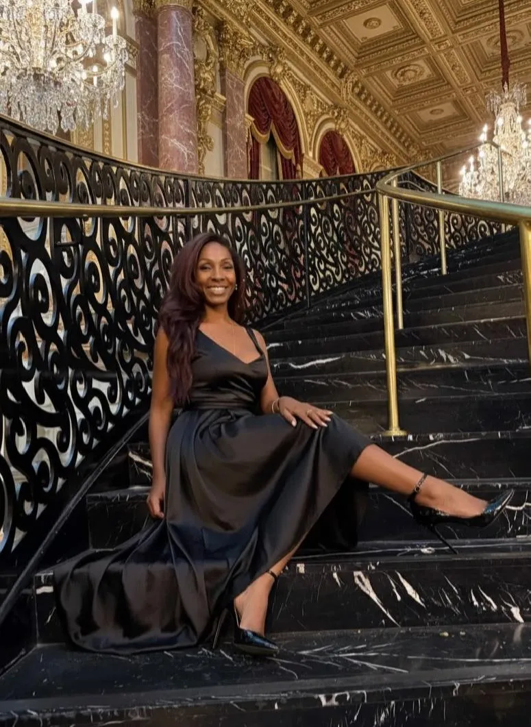 Woman in a black dress sitting on a marble staircase with ornate railings and chandeliers in an elegant interior.