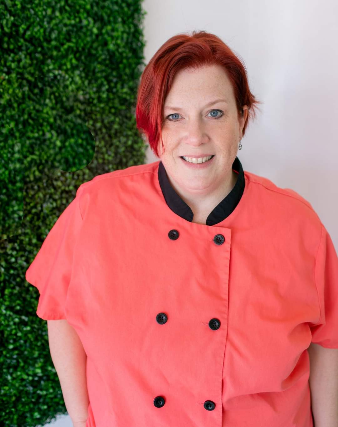 Professional headshot of pastry chef Erica Bentson in a coral chef coat against a green hedge background