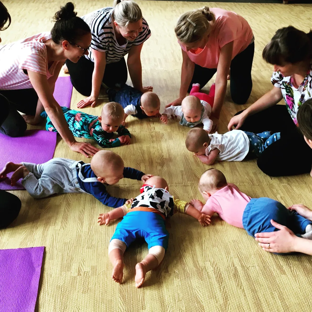 Mothers and babies lying in a circle on yoga mats, smiling and engaging in a family yoga class together.