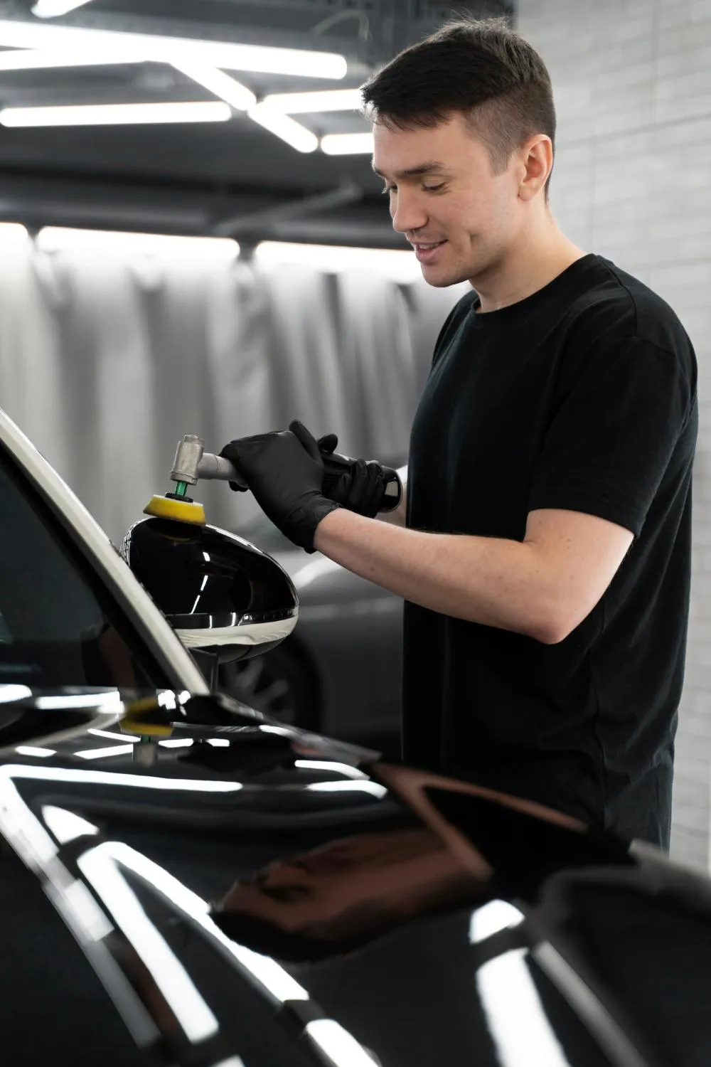 Young man in auto shop using a buffer to polish the side mirror of a black car under restoration