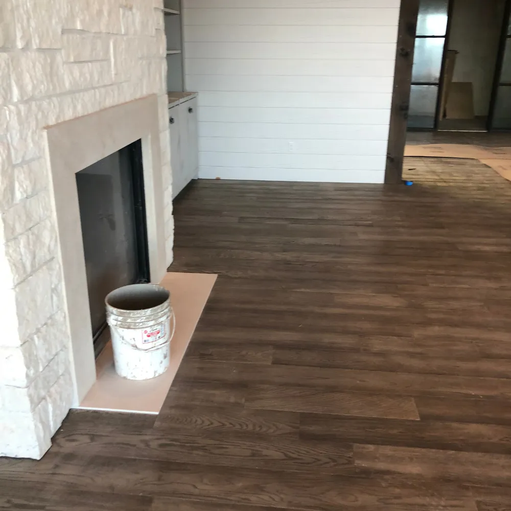 Living room with rich dark wood flooring, a stone fireplace, and white shiplap walls, partially staged with a paint bucket in the foreground.