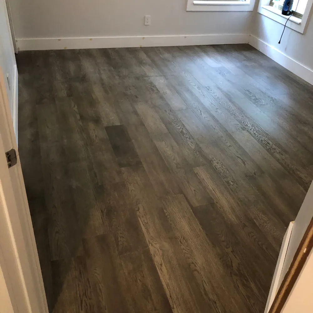 Empty room featuring gray-toned wood flooring, clean white baseboards, and natural light streaming through corner windows.