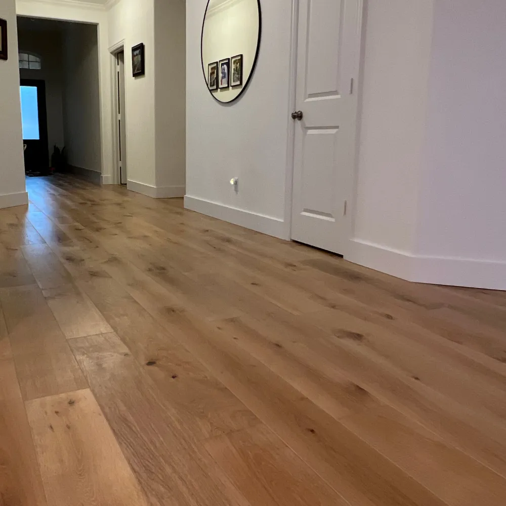 Hallway with light natural wood flooring, white walls, a circular mirror, and framed photos, leading to a well-lit entryway.