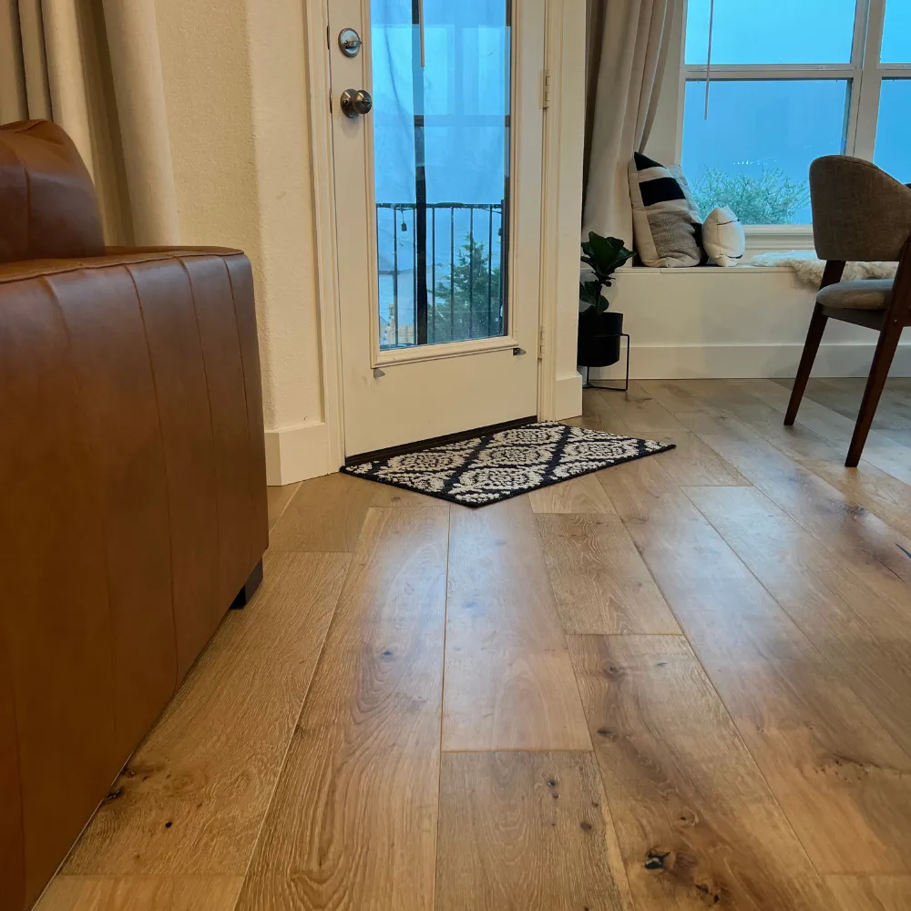 Cozy living space with light natural wood flooring, a leather sofa, a small black-and-white patterned rug by a glass door, and a seating nook with pillows and a view of an outdoor balcony.