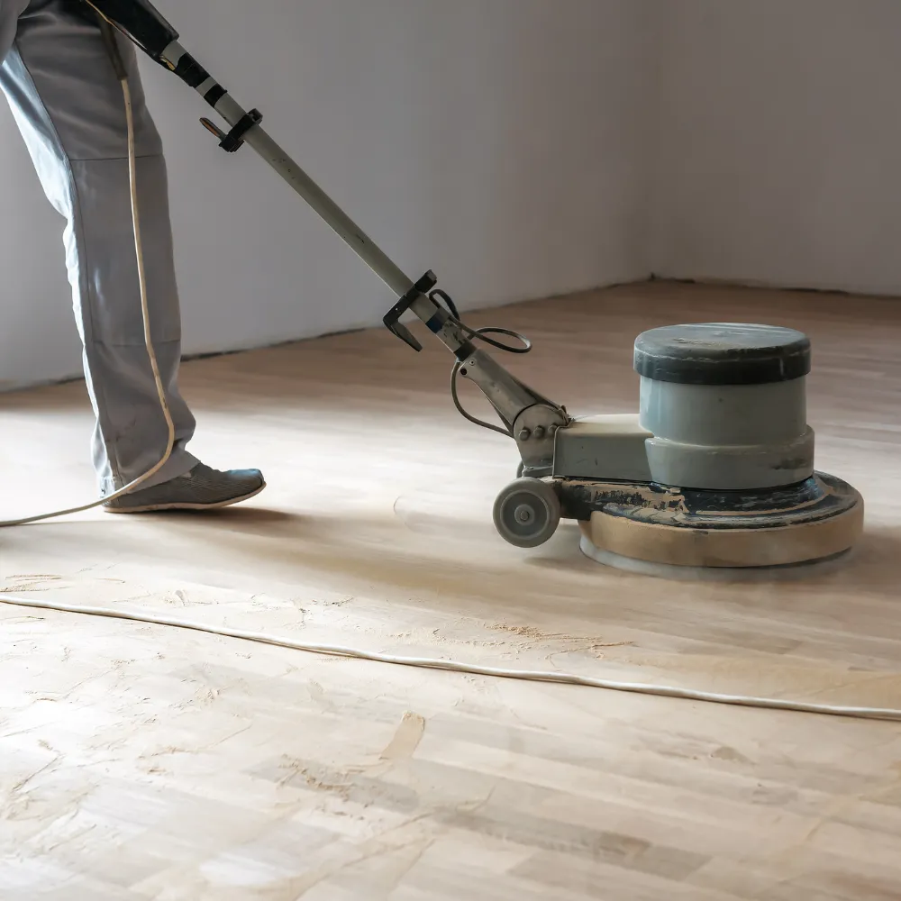 Worker using a floor buffer to sand and smooth a hardwood floor, preparing it for finishing in an empty room.