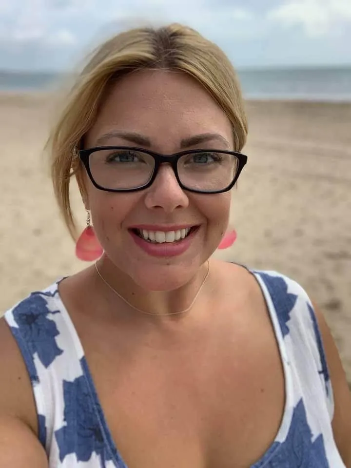 Nikki, a smiling woman with blonde hair wearing glasses and a sleeveless top, standing on a beach with the ocean in the background