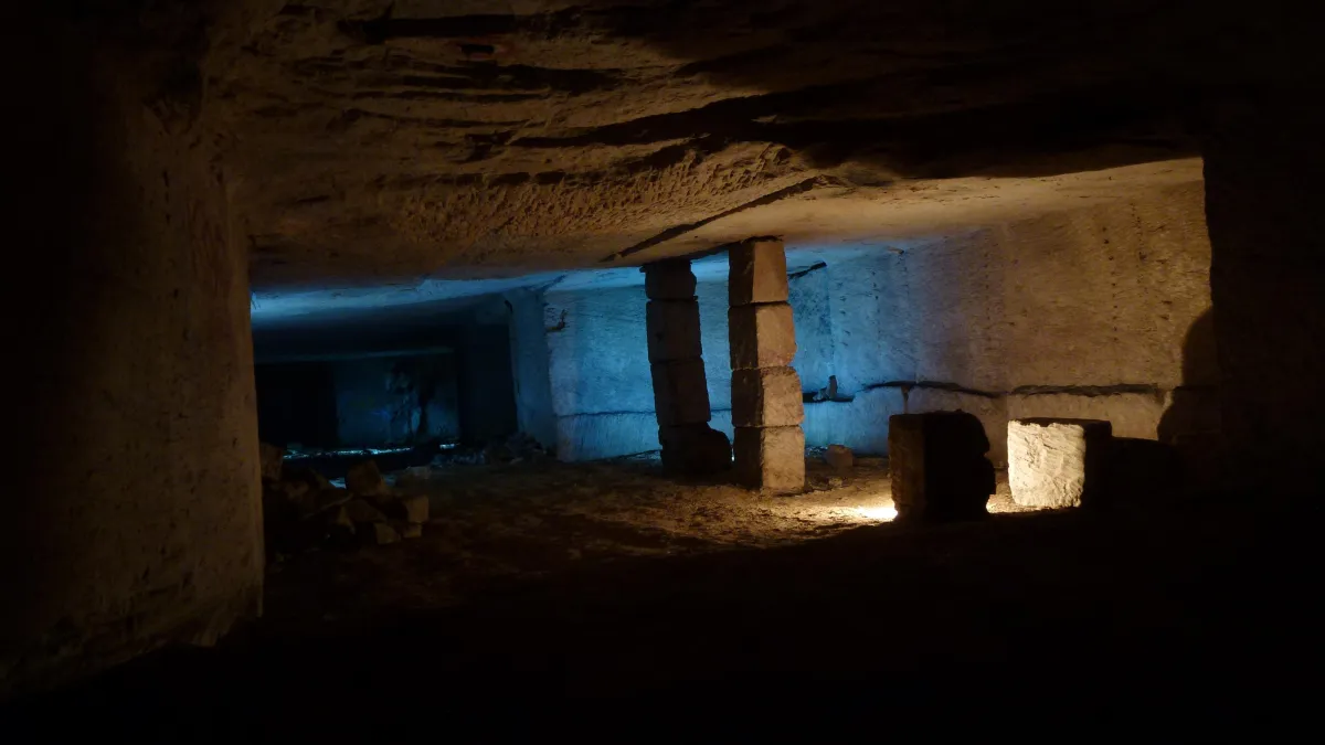 An underground tunnel or cave chamber dimly lit with blue and yellow lighting. The space has rough, chiseled stone walls and ceiling, with stacked stone pillars supporting the structure. Rubble and debris are scattered across the dirt floor, giving it an abandoned or archaeological feel.
