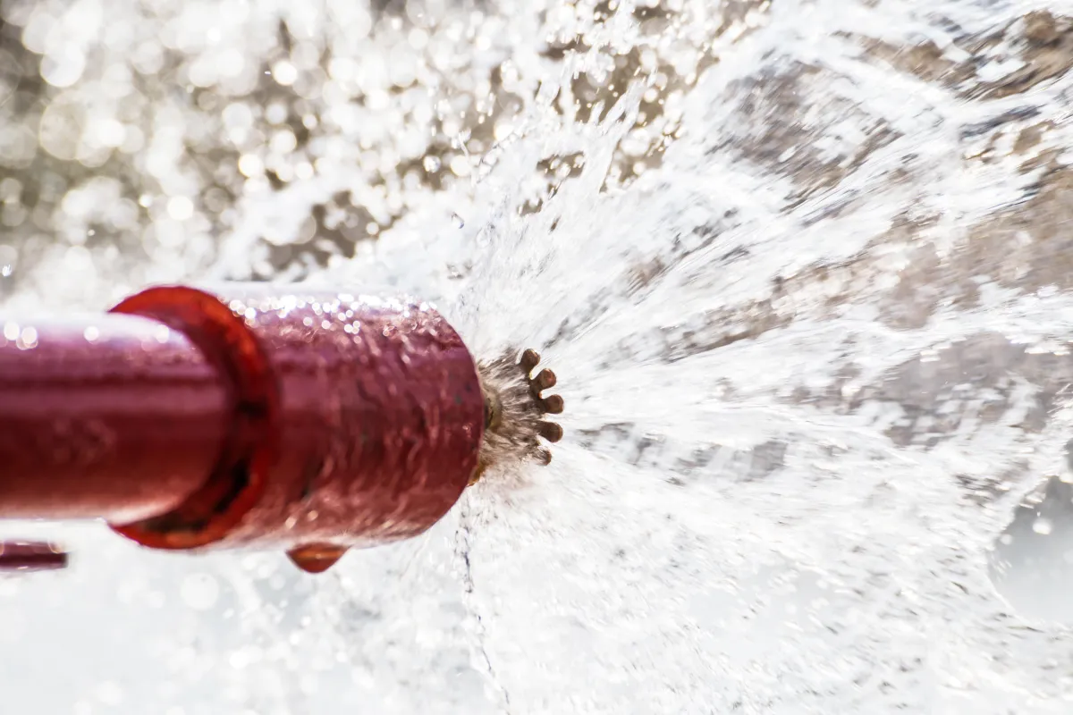 A close-up of a red pipe with a major leak, spraying water forcefully from a cracked joint. The high-pressure burst creates a dramatic spray, highlighting a significant plumbing failure or emergency.
