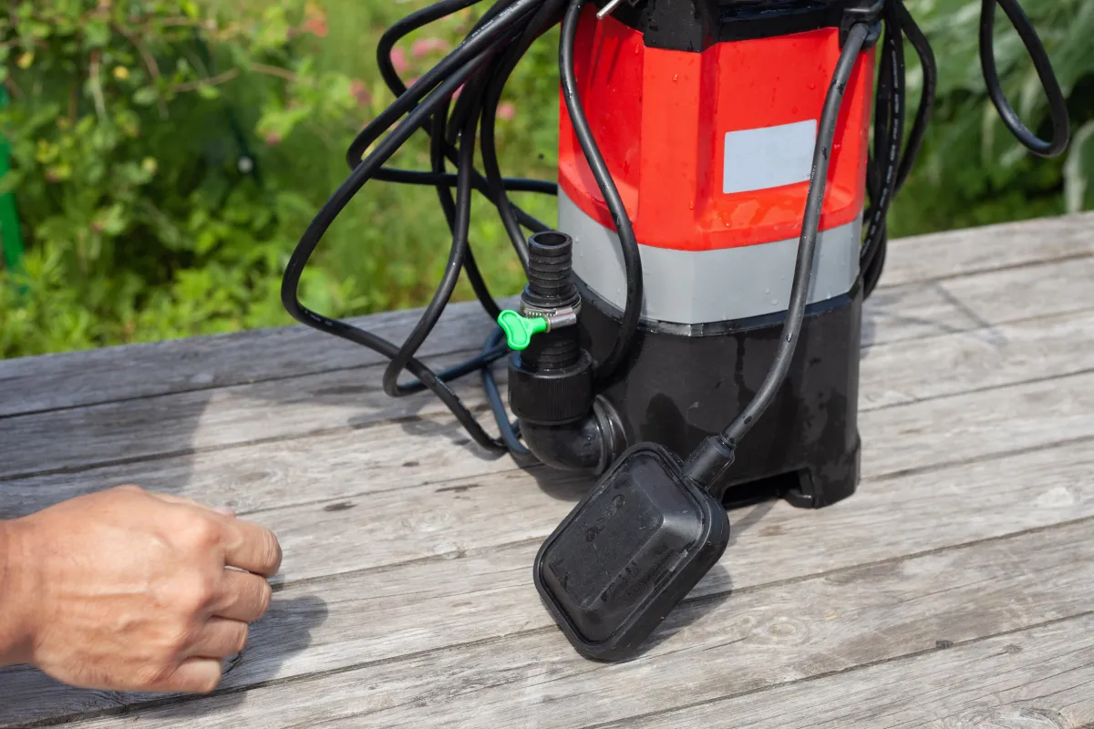 A submersible water pump with a red and black body sits on a wooden surface, surrounded by electrical cords and a float switch. A person’s hand is partially visible near the pump, suggesting setup or inspection. The background includes greenery, indicating an outdoor setting.
