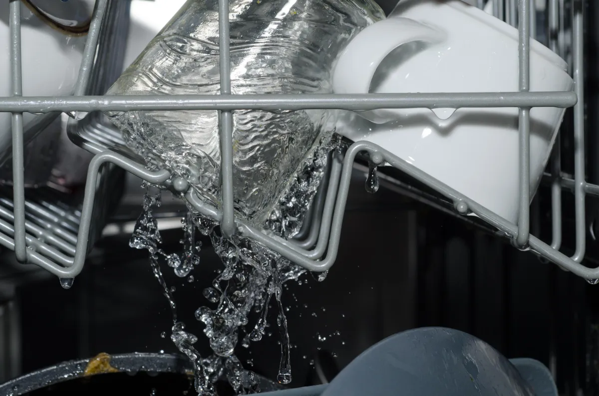 Close-up view of water spraying inside a dishwasher during a wash cycle. A glass and white ceramic mug sit on the top rack, with droplets and streams of water splashing around the dishes, indicating the machine is actively running.