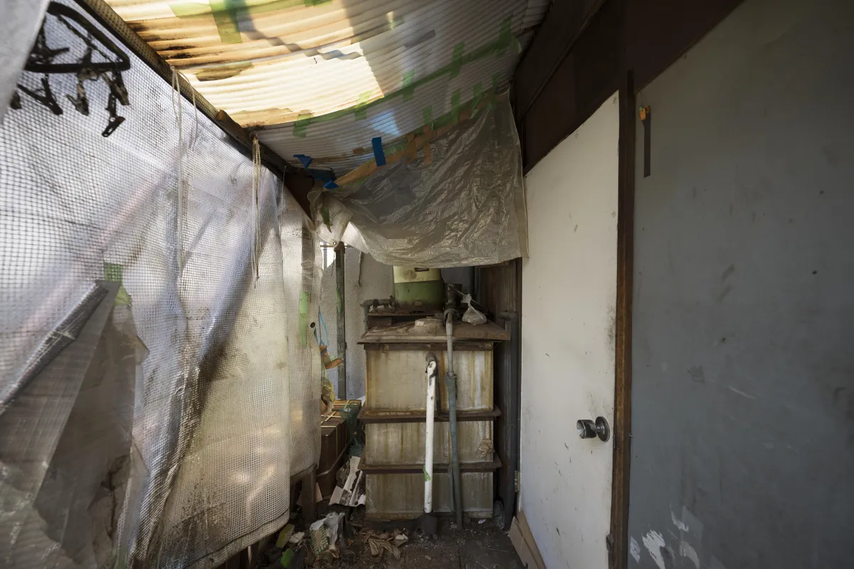 A narrow, cluttered utility area with makeshift plastic sheeting taped to the ceiling and walls, likely used for temporary weather protection. Pipes and an old water system are visible, along with a closed white door on the right. The space appears worn and in need of repair or maintenance.