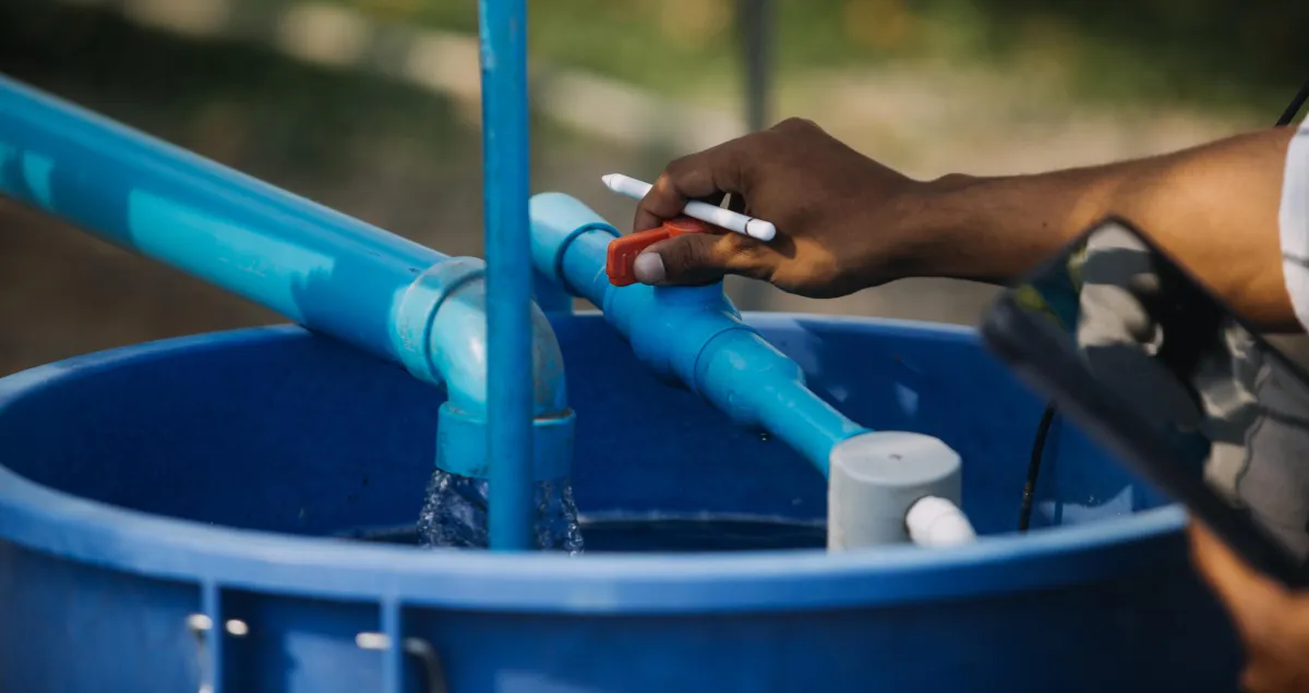 A close-up of a person adjusting a red valve on a blue PVC pipe system connected to a large blue container. Water is visibly flowing through the system, and the person is holding a pen and a tablet, possibly performing a system check or water quality test.