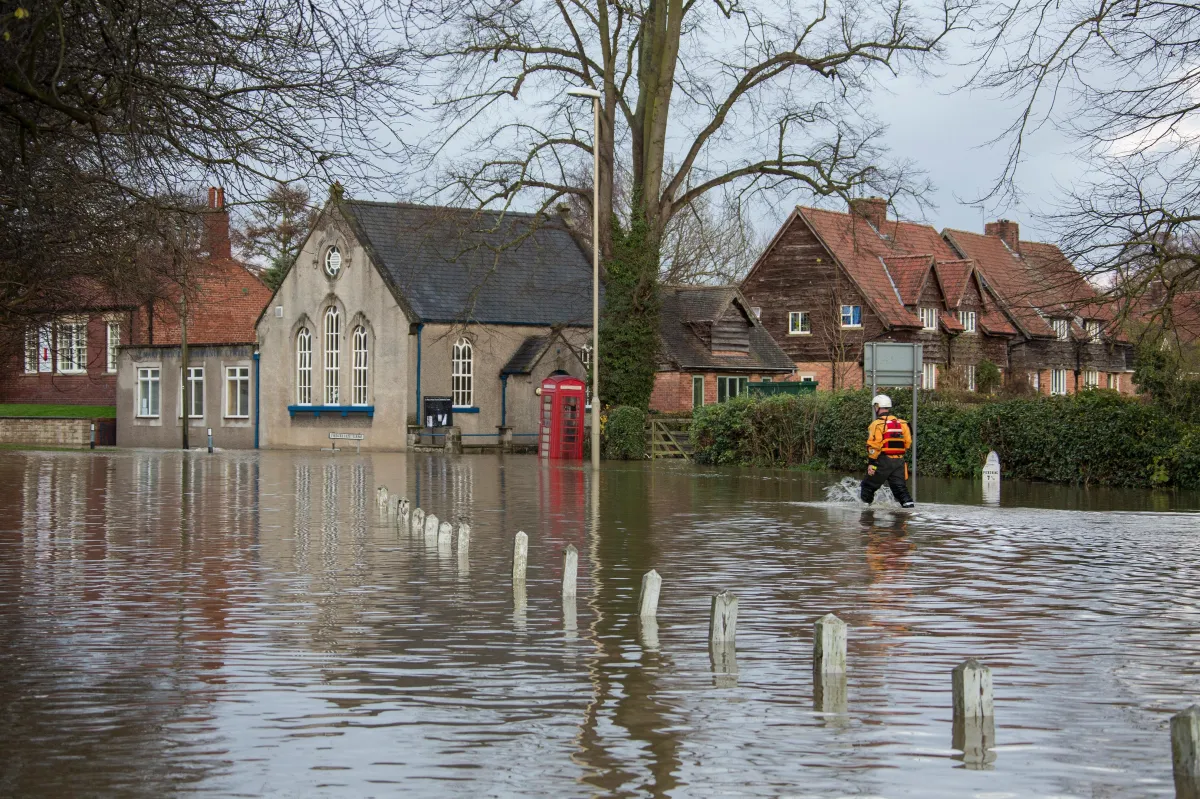 A flooded residential street in a village, with water submerging the road up to the doorsteps of houses and a historic stone building. A person in safety gear wades through the floodwaters, and a red phone booth is partially submerged near the building. Trees without leaves suggest it is winter or early spring.