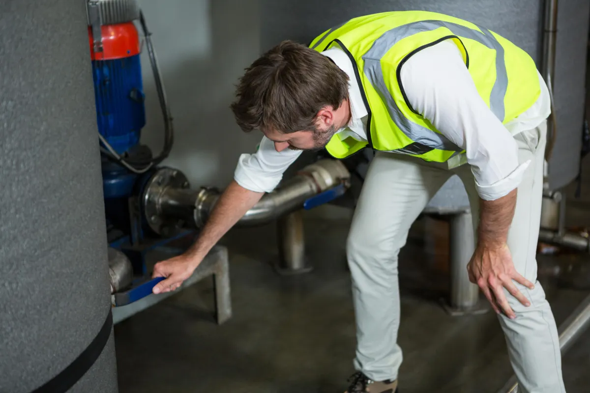A maintenance worker wearing a high-visibility safety vest and white pants inspects a pipe connection in an industrial facility. He is leaning forward, closely examining a valve or fitting attached to large insulated tanks and metal piping.
